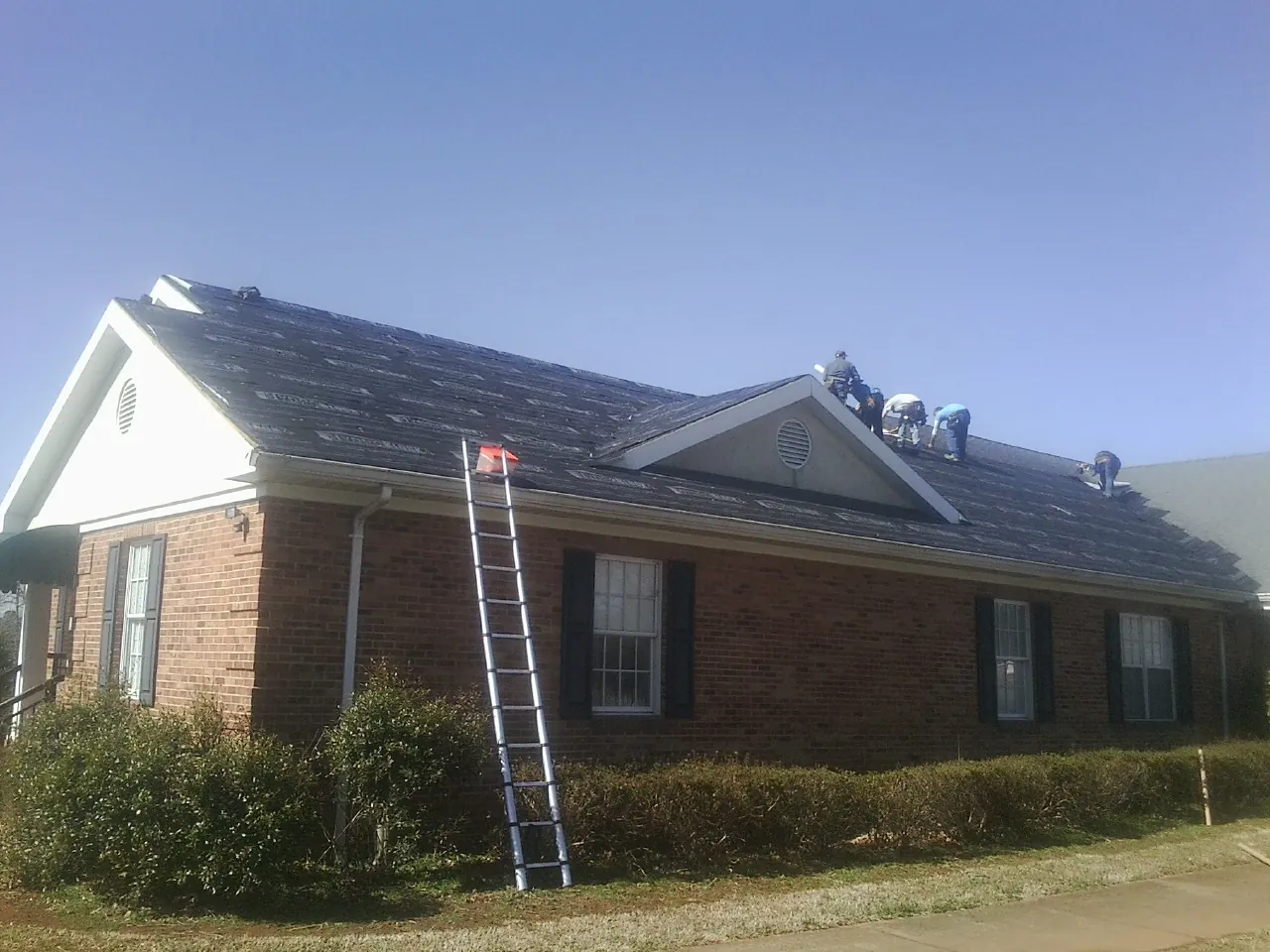 Crew tearing shingles off a brick ranch roof with a ladder set — Emergency Roof Repair work by our crew in Janesville